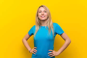 Obraz premium Young blonde woman over isolated yellow background posing with arms at hip and smiling