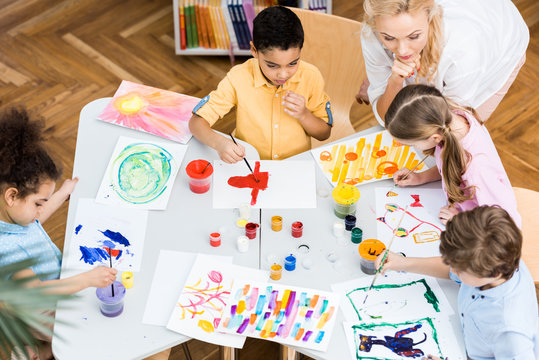 overhead view of woman looking at multicultural kids painting on papers