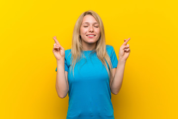 Young blonde woman over isolated yellow background with fingers crossing and wishing the best