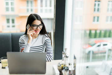Fototapeta premium Young smiling businesswoman sitting at table in coffee shop and uses smartphone on table cup of coffee and notebook