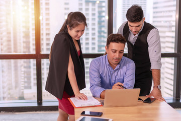 Group businesspeople colleagues talking about planning project in the workplace. Man holding pen with colleagues sitting on desk with laptop while talking work together in office on city background.