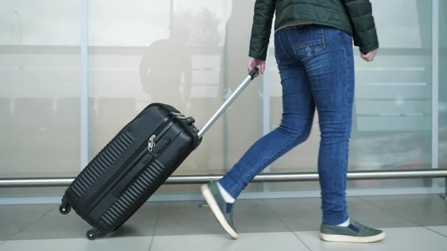 Traveler woman in jeans and lacket is walking in modern airport terminal carrying suitcase, side view. Passenger is going to her flight in city airport, closeup legs.