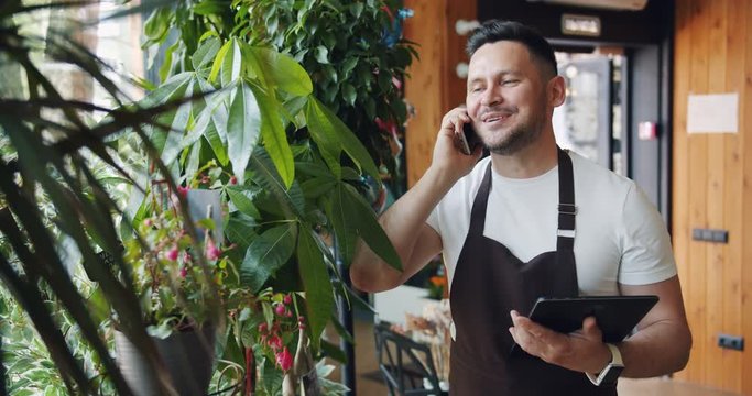Joyful owner of florist shop speaking on mobile phone smling holding tablet standing on store alone looking at flowers. People and communication concept.