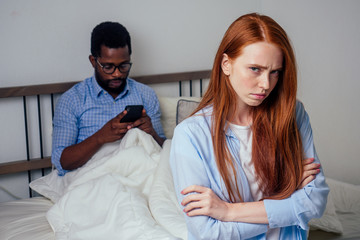 Fototapeta premium redhaired ginger caucasian happy female and multi-ethnic afro man together lying in bed bedroom looking at smartphones texting.Family conflict