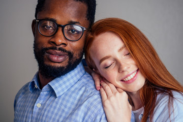 young redhead freckled woman hugging her African boyfriend, looking at the camera with happy face expression