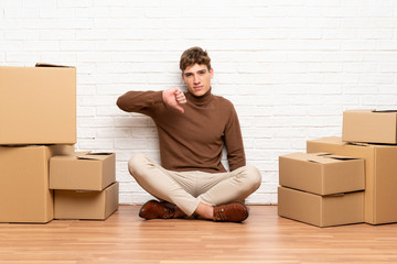 Handsome young man moving in new home among boxes showing thumb down sign