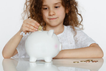 Cute curly little girl putting coin into big white piggy bank, over white background. Educational, saving money concept.