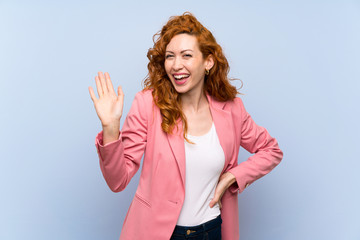 Redhead woman in suit over isolated blue wall saluting with hand with happy expression