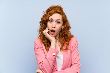 Redhead woman in suit over isolated blue wall surprised and shocked while looking right