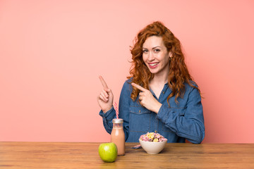 Redhead woman having breakfast cereals and fruit pointing finger to the side