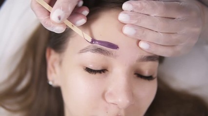 Young, caucasian girl is lying on couch during eyebrowes treatment at studio beauty, beautician depilating and shaping brows at beauty salon. Master applying a line of wax above the brow using a stick - Powered by Adobe