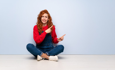 Redhead woman with overalls sitting on the floor frightened and pointing to the side