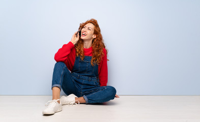 Redhead woman with overalls sitting on the floor keeping a conversation with the mobile phone