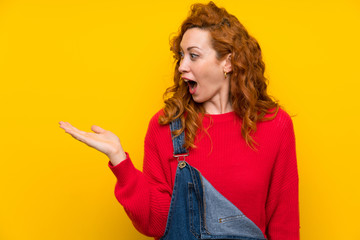 Redhead woman with overalls over isolated yellow wall holding copyspace imaginary on the palm