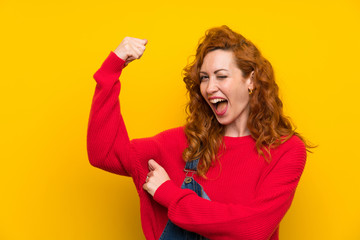 Redhead woman with overalls over isolated yellow wall making strong gesture