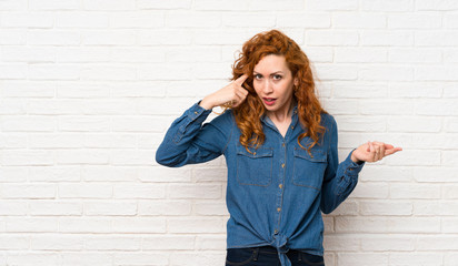 Redhead woman over white brick wall making the gesture of madness putting finger on the head