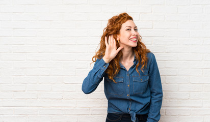 Redhead woman over white brick wall listening to something by putting hand on the ear