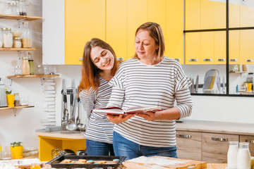 Family leisure in kitchen. Mother and daughter enjoying cooking together, comparing result with recipe in cookery book.