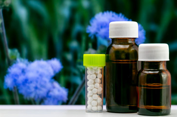 Advanced Homeopathic Concept - Close up image of homeopathic globule sugar pills and liquid substance in glass bottle with blurred blue flower and green leaves background