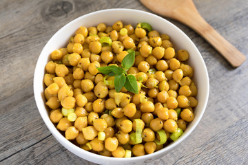 Cooked chickpeas with leek, chia seeds, sesame seeds and basil on top in white bowl. Wooden kitchen surface with wooden ladle.