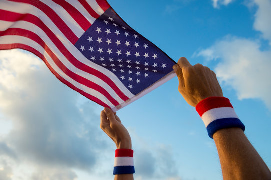 Hand with USA red, white, and blue wristbands holding an American flag waving in bright sunny blue sky