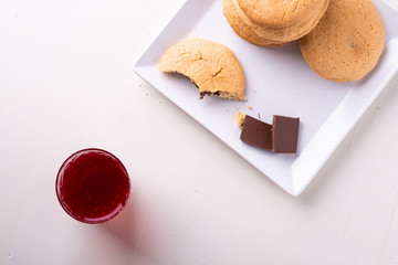 Homemade stack of shortbread chocolate cookies with one bitten cookie and two pieces of chocolate on white plate with stewed fruit red compote drink in glass on wooden table top view