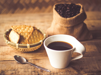 Black coffee In the cup white have roasted coffee beans are In a cloth bag sack butter cracker In the basket weave bamboo, and still have stainless steel spoon all placed on a wooden table.