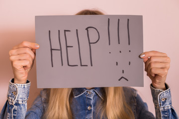 Woman holding paper with word help and sad face while standing in front of the wall.