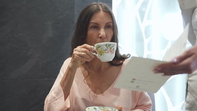 Charming and calm woman spending free time in bright and light spa salon with modern interior. Female drinking beverage and listen information about new procedure in resort center