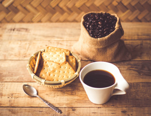 Black coffee In the cup white have roasted coffee beans are In a cloth bag sack butter cracker In the basket weave bamboo, and still have stainless steel spoon all placed on a wooden table.