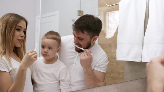 Waist-up shot of lovely Caucasian family, dressed in white t-shirts, with 3-year-old son standing together in front of mirror in bathroom, and little boy watching while his parents are brushing teeth