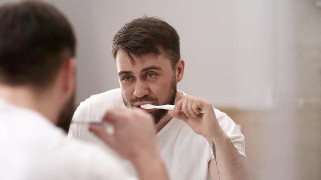Over-shoulder shot of handsome bearded Caucasian man in white t-shirt brushing his teeth with toothpaste in bathroom and looking at his reflection in mirror