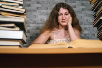 Tired female student reading among books. Pensive young woman sitting at table with pile of book and reading on background of grey wall