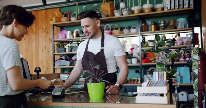 Young woman customer is buying beautiful houseplant in pot paying with credit card talking to male cashier smiling. Business, payment and people concept.