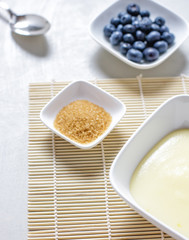Vanillepudding, Blaubeeren, brauner Zucker in weißen Servierschalen auf weißem Holztisch, Ansicht...