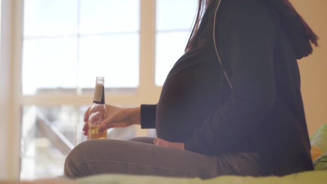 Unrecognizable young pregnant woman drinking beer alone sitting on the sofa at home. Unhealthy lifestyle. Bad habit. The concept of unhealthy motherhood.