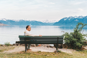 Naklejka na ściany i meble Portrait of handsome man with sitting on bench, meditating by the lake, wearing white sweatshirt