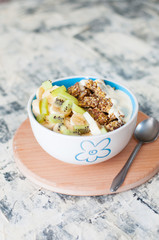 Greek yogurt, granola, kiwi and banana in bowl with a spoon on a gray concrete background, wooden tray, vertical format. Fitness diet for weight loss, proper and tasty food