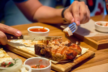 A piece of kebab, on a wooden plate, men's hands holding a fork and a knife