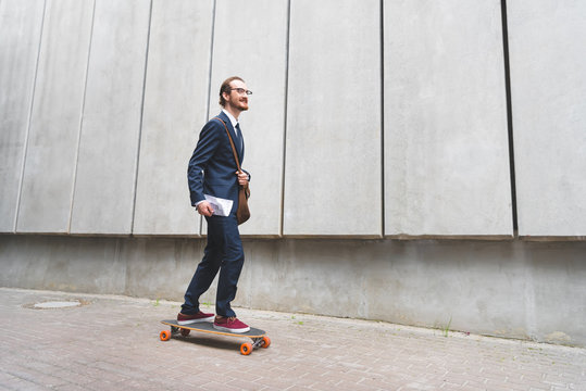 Happy businessman in formal wear riding on skateboard, looking away