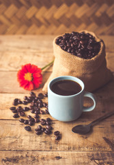Black coffee In the cup sky blue color have roasted coffee beans are In a cloth bag sack and on table with, still have red flowers and stainless steel spoon all placed on a wooden table.