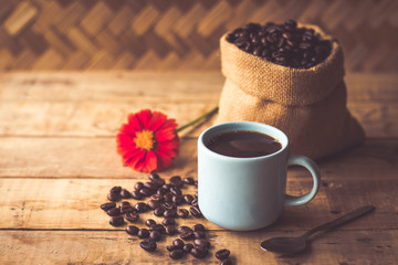 Black coffee In the cup sky blue color have roasted coffee beans are In a cloth bag sack and on table with, still have red flowers and stainless steel spoon all placed on a wooden table.