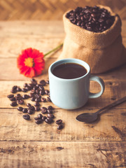 Black coffee In the cup sky blue color have roasted coffee beans are In a cloth bag sack and on table with, still have red flowers and stainless steel spoon all placed on a wooden table.