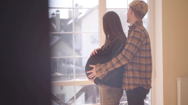 Teenage couple at home together. The young man caressing the belly of his pregnant girlfriend standing behind her. A minor girl carries a child. Slow motion.