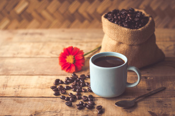 Black coffee In the cup sky blue color have roasted coffee beans are In a cloth bag sack and on table with, still have red flowers and stainless steel spoon all placed on a wooden table.