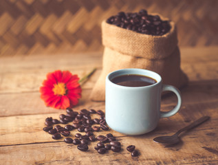 Black coffee In the cup sky blue color have roasted coffee beans are In a cloth bag sack and on table with, still have red flowers and stainless steel spoon all placed on a wooden table.