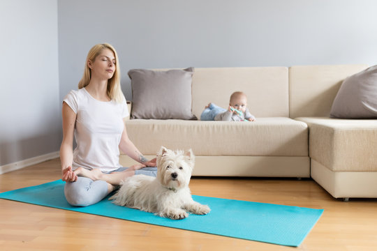 Mother and child with white dog in a bright room with grey walls and wooden floor on blue mat exercising yoga calm fitness white sofa counch in home 