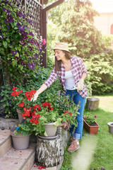 Beautiful smiling Caucasian brunette with hat, gardening gloves and in working clothes taking care of her flowers in backyard.