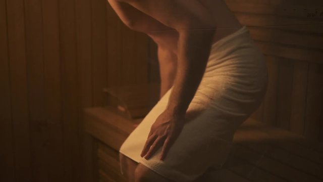 Beautiful handsome young man is sitting on wooden bench at sauna in white towel. Strong sexy and healthy body is sweating from heat in traditional steam room. Spa wellness and wellbeing therapy. Calm