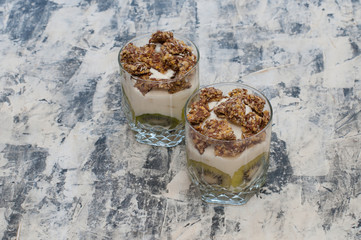 Granola, kiwi, banana and greek yogurt in glass cups on gray concrete
